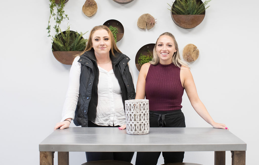 Two women standing at table with plants hanging on wall behind them.