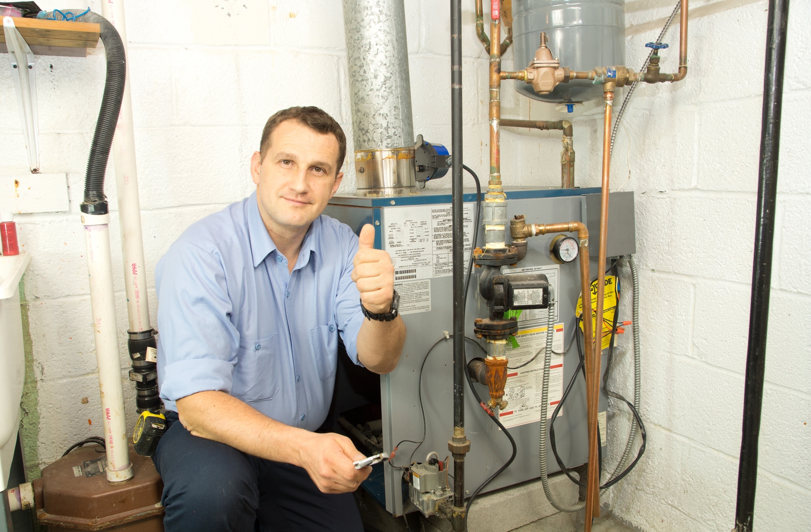 Man giving thumbs up while sitting beside furnace in basement of house