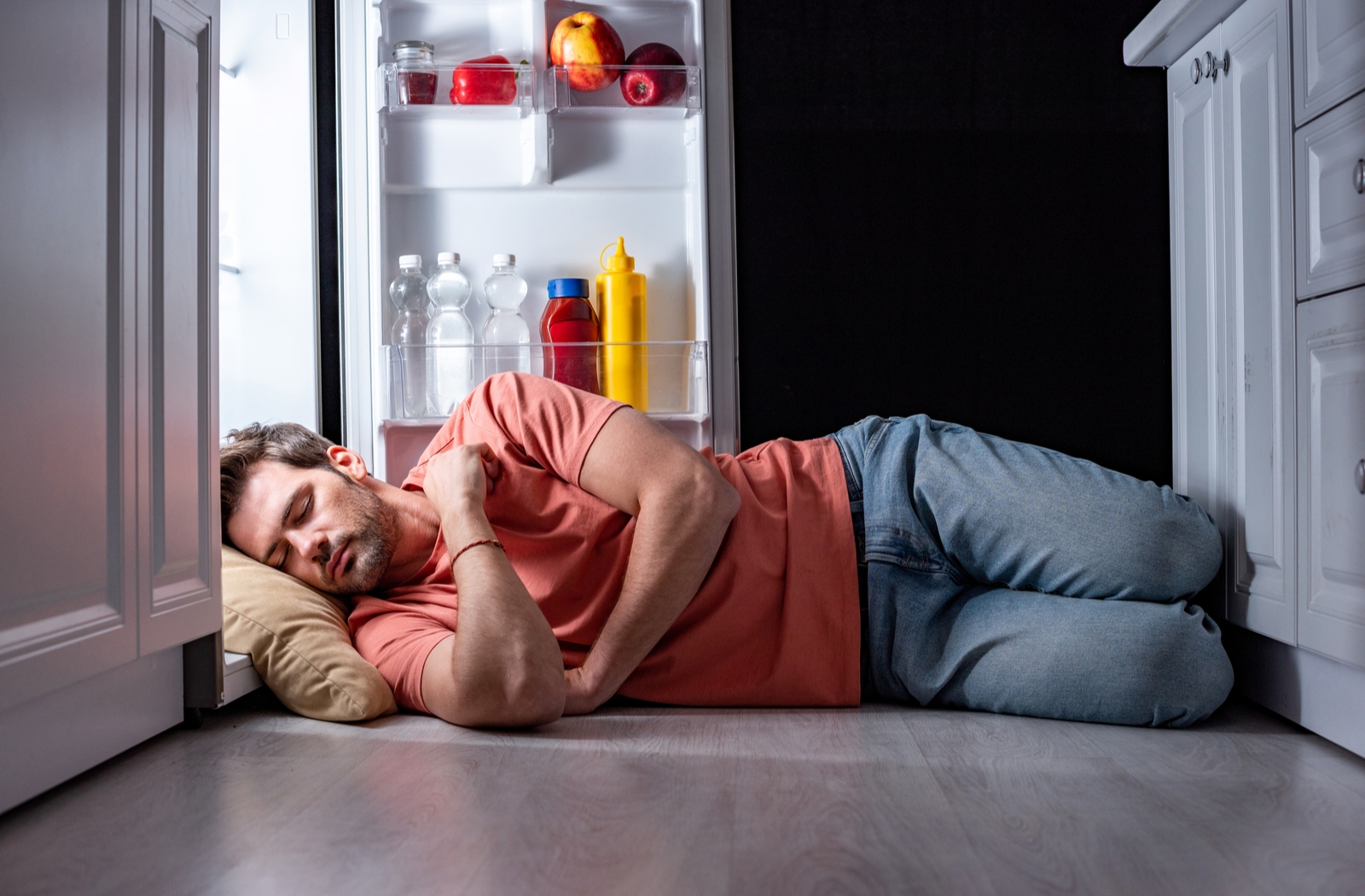 Man sleeping on the floor in the kitchen in front of an open refrigerator because his house is too warm to sleep in the summer