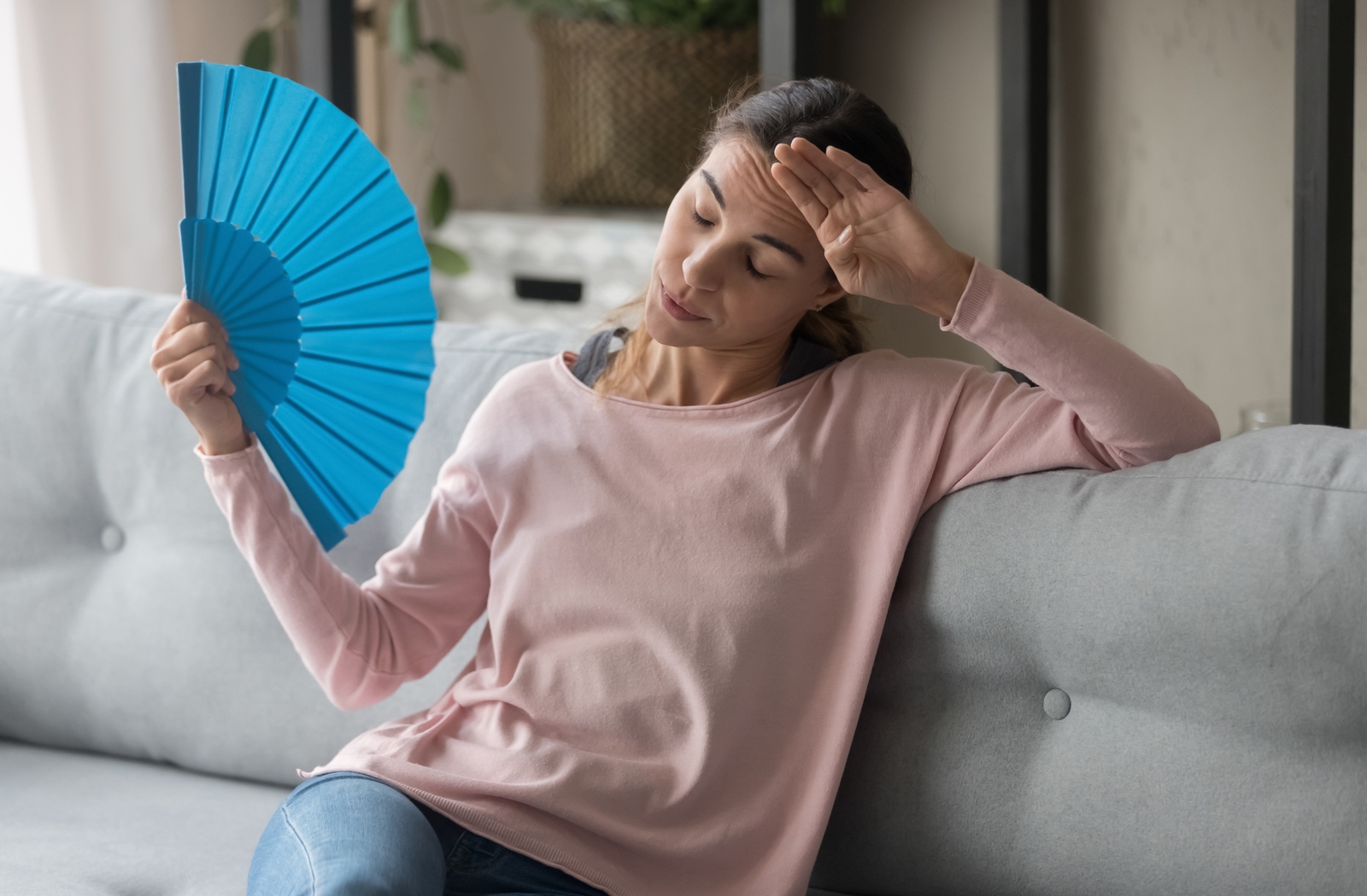 Woman sitting on her couch overheating using a paper fan to try to cool herself down because her air conditioning failed