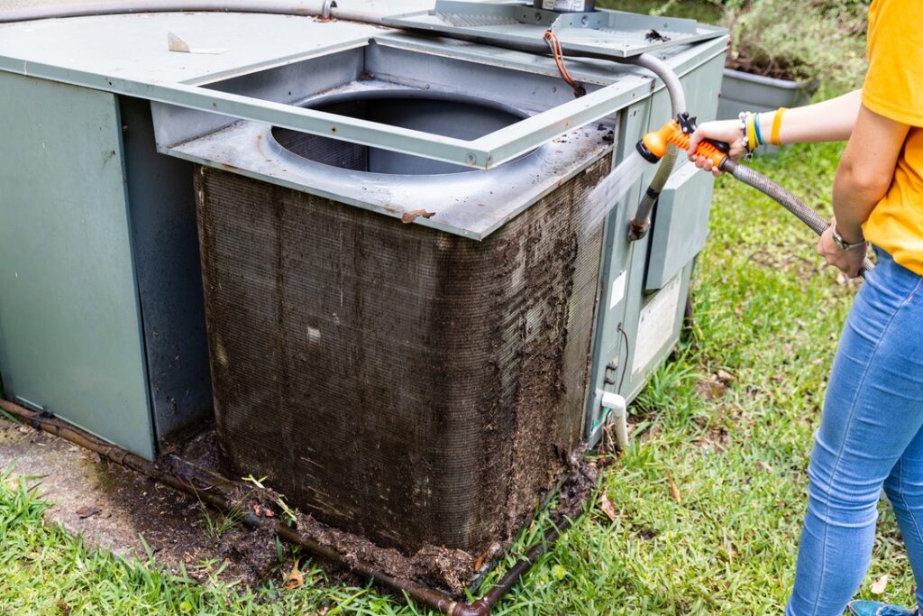 Person cleaning outdoor AC unit with hose before winter