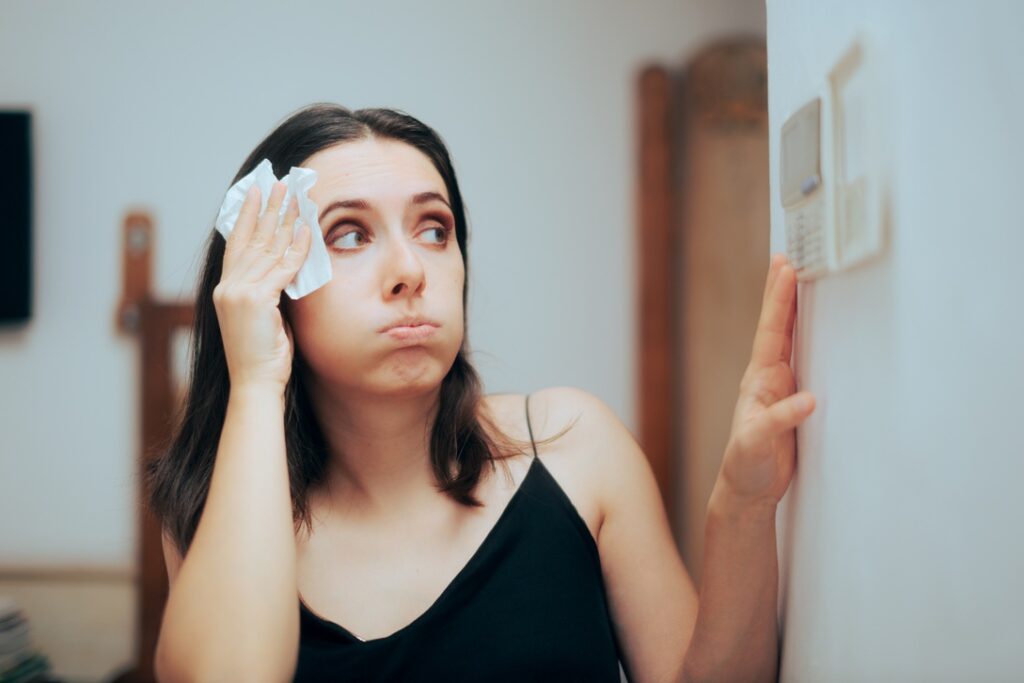Woman sweating and wiping forehead with cloth while setting thermostat during summer