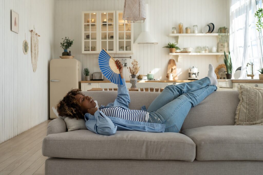 Woman lying on couch and fanning self to keep cool to demonstrate the importance of correct thermostat settings