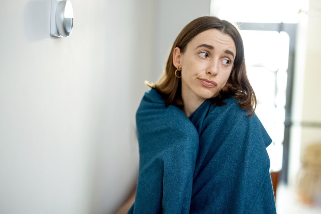 Woman wearing blankets near thermostat to represent failure to start furnace in winter