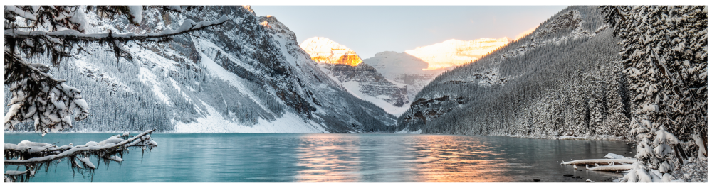 A serene winter landscape showcasing the cold, intense weather of Alberta, with a snow-covered forest surrounding a turquoise lake.