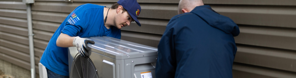 Two Action Furnace Technician's carefully install a heat pump on the side of a house.